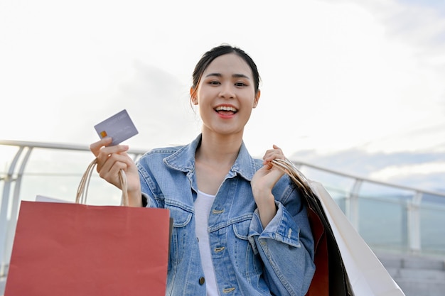 Aanbiddelijke Aziatische vrouw die creditcard vasthoudt en boodschappentassen draagt