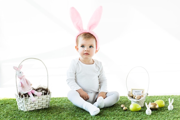 Aanbiddelijke baby in de hoofdband van konijnenoren die dichtbij stromanden zit met kleurrijke paaseieren en