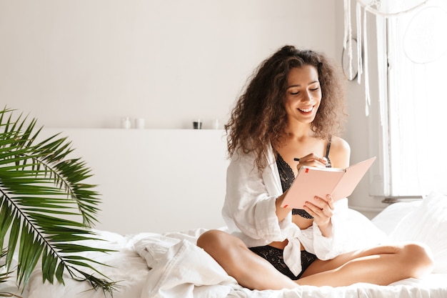 aanbiddelijke jonge vrouw die lingerie draagt die lacht en dagboekboek op bed in wit appartement houdt