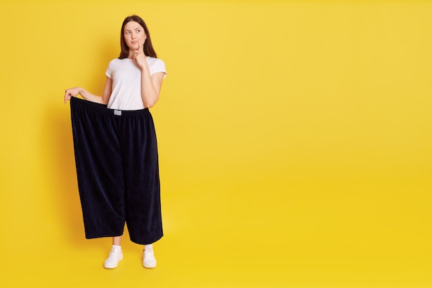 Aanbiddelijke vrouw die een grote broek draagt die zich met doordachte gezichtsuitdrukking bevindt, de vinger op de lip houdt, wegkijkend, ruimte voor promotie kopieert, geïsoleerd over gele muur.