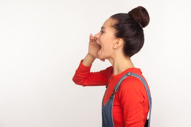 Aandacht, aankondiging. Zijaanzicht van stijlvol meisje met haarbroodje in denim overall met arm in de buurt van wijd open mond en schreeuwende boodschap, luide reclame. studio-opname geïsoleerd op een witte achtergrond