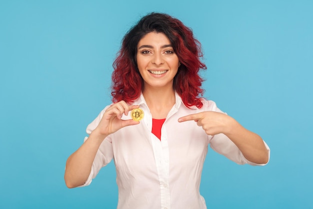 Aandacht voor cryptocurrency Portret van charmante vrouw met mooi rood haar die bitcoin wijst en glimlacht naar de camera die digitaal geld elektronische handel binnen studio opname blauwe achtergrond toont