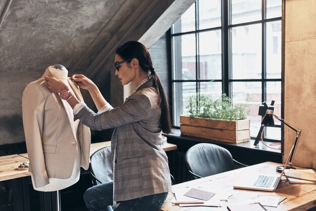 Aandacht voor elk detail. Ernstige jonge vrouw in brillen die een kraag van de jas op de mannequin aanpast terwijl ze in haar atelier staat