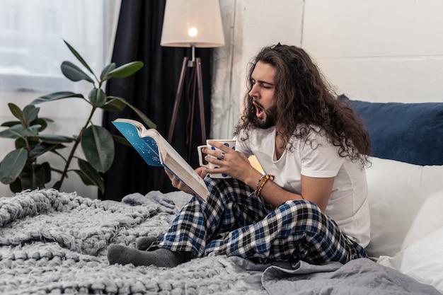 Aangenaam drankje. Aardige jonge man die een kopje thee vasthoudt terwijl hij met een boek op zijn bed zit