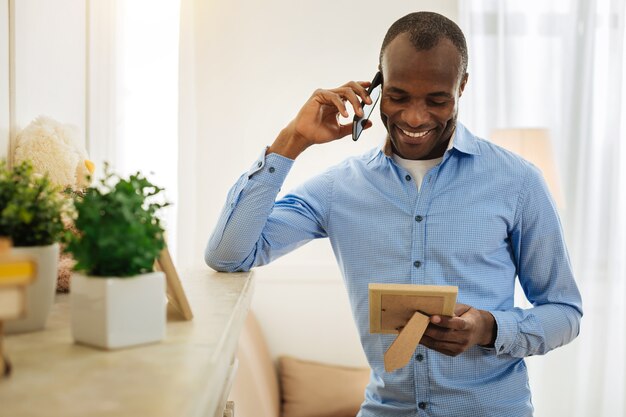 Aangenaam gesprek. Knappe geïnspireerde jongeman praten aan de telefoon en glimlachen terwijl vasthouden en kijken naar de foto