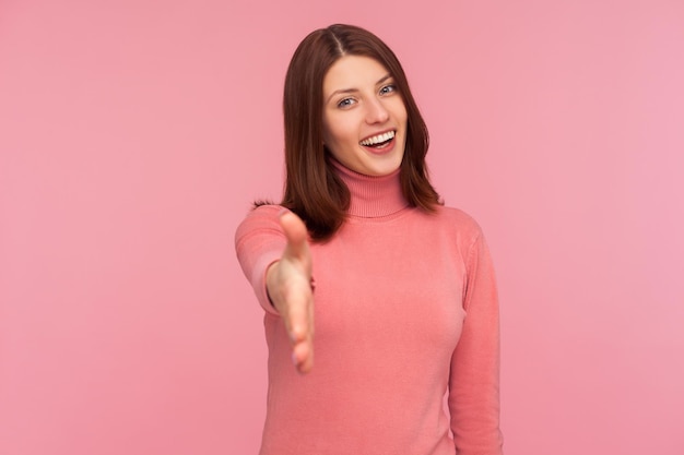 Aangenaam je te ontmoeten Vriendelijke lachende vrouw in roze trui die haar hand uitstrekt om gasten te verwelkomen gastvrijheid Indoor studio-opname geïsoleerd op roze achtergrond