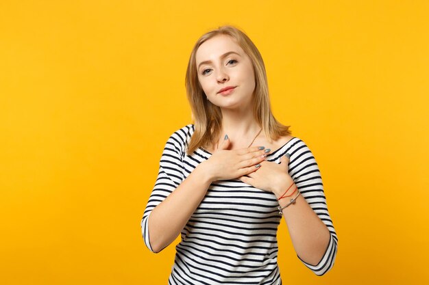 Aangename jonge vrouw in gestreepte kleding op zoek naar camera, hand in hand gekruist op hart, borst geïsoleerd op geeloranje muur achtergrond. Mensen oprechte emoties levensstijl concept. Bespotten kopie ruimte.