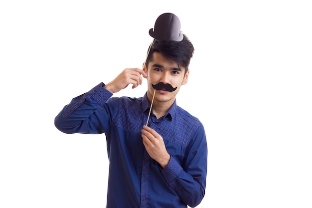 Aangename jongeman met donker haar in blauw shirt met kartonnen snorren en zwarte hoed op een witte achtergrond in de studio