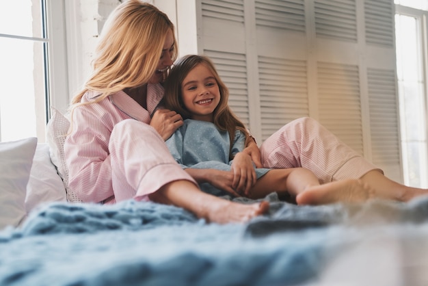 Aangename ochtend. Mooie jonge moeder die haar dochter omhelst en glimlacht terwijl ze thuis op bed zit