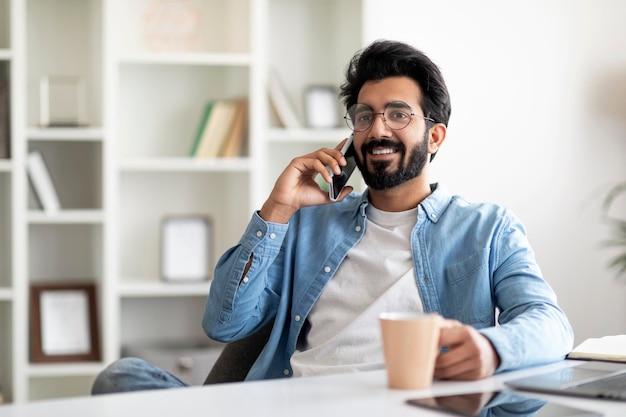 Aangename oproep lachende knappe indiase man praten op mobiele telefoon en koffie drinken