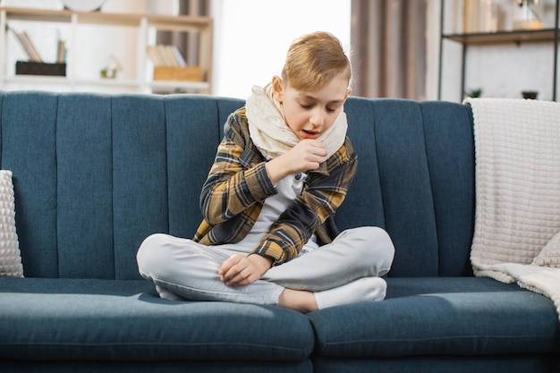 Aangename vermoeide zieke tienerjongen met sjaal om de nek zittend op een zachte bank en hoestend