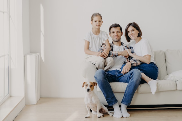 Aanhankelijk familie poseren samen op de bank in lege ruime kamer met witte muren, hun favoriete hond zit op de vloer