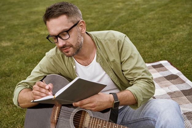 Aantekeningen schrijven jonge gefocuste man met een bril die akoestische gitaar vasthoudt en een lied componeert