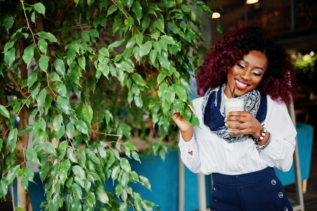 Aantrekkelijk afrikaans-amerikaans krullend meisje in witte blouse en blauwe korte broek geposeerd in café tegen boom met latte bij de hand