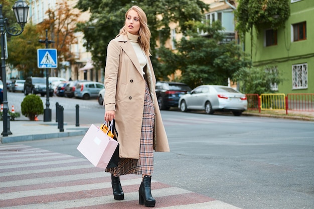 Aantrekkelijk, casual blond meisje in stijlvolle trenchcoat met boodschappentassen die bedachtzaam op zebrapad lopen