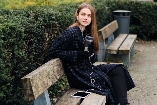 Aantrekkelijk casual meisje met thermokop die muziek luistert in oortelefoons die op een bankje in het stadspark rusten