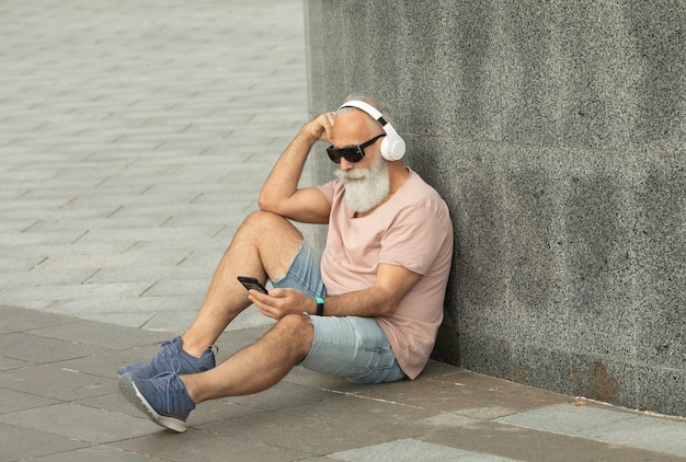 Aantrekkelijk en glimlachend bebaarde oude senior man met wit haar genieten van muziek van slimme telefoon buiten dragen witte koptelefoon.