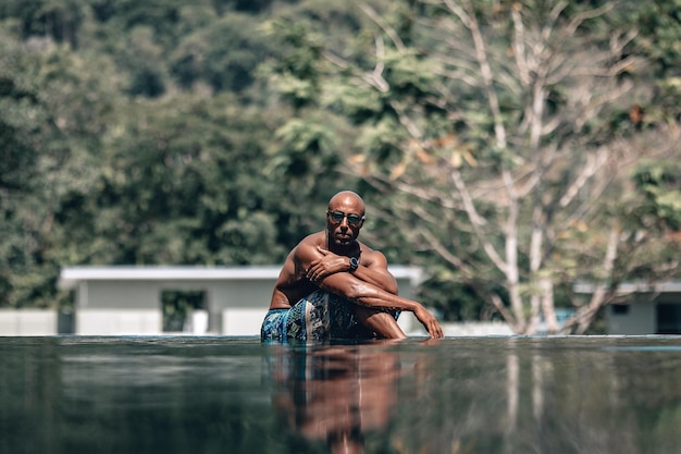 Aantrekkelijk en ontspannen Arabisch shirtloos mannetje in turquoise sarong liggend aan de zijkant van een overloopzwembad in een zonnige dag; reflectie in het water, palmen en hotel op de achtergrond; vakantie concept.