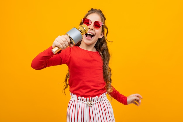 Aantrekkelijk Europees meisje dat in zonnebril met een microfoon op een gele achtergrond zingt