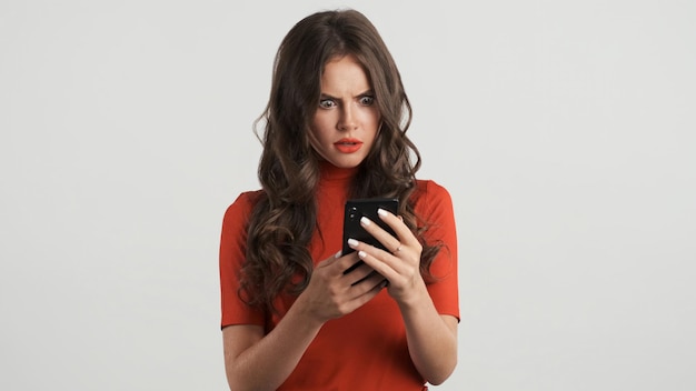 Aantrekkelijk geschokt brunette meisje in rode top verbaasd met behulp van smartphone over grijze achtergrond