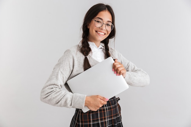 Aantrekkelijk glimlachend schoolmeisje met een unifrom die over een witte muur staat en een laptop draagt