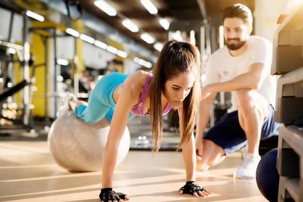 Aantrekkelijk jong atletisch meisje doet push-ups met fitness bal in de moderne sportschool naast de knappe tevreden personal trainer.