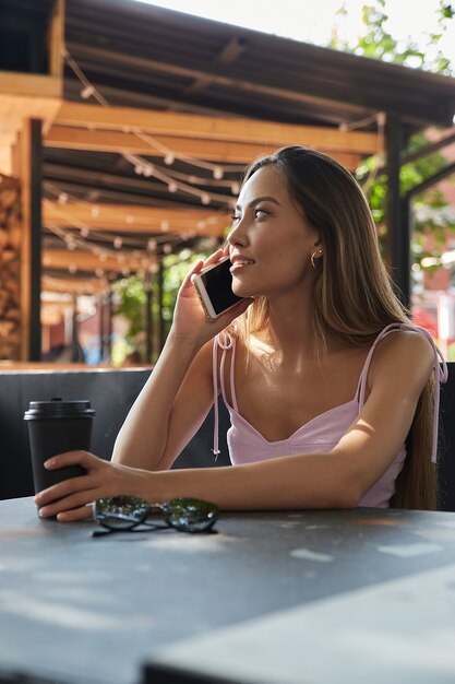 Aantrekkelijk jong aziatisch meisje in roze jurk bellen door smartphone koffie drinken