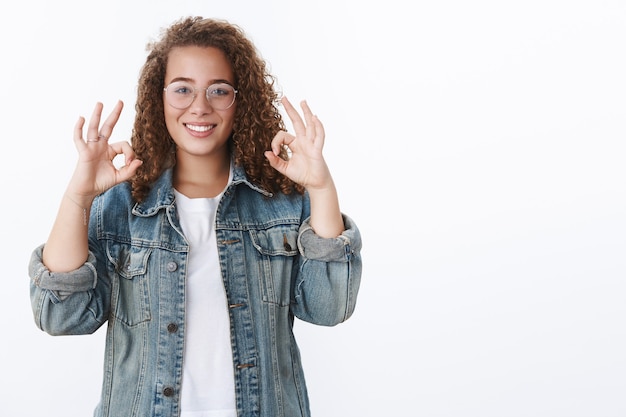 Aantrekkelijk jong freelance meisje met krullend haar toont goedkeuring oke ok gebaar glimlachend tevreden eens concept interessant ding idee uitstekend, opgetogen witte muur