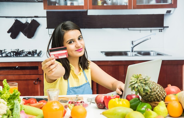 Aantrekkelijk jong Indiaas meisje met schort in keuken online winkelen met bankpas of creditcard op laptop met tafel vol met verse groenten en fruit