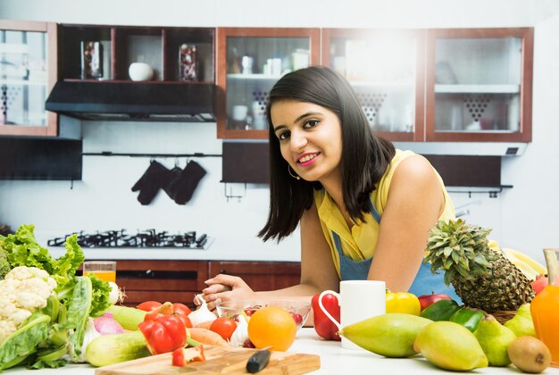 Aantrekkelijk jong Indiaas meisje met schort koken in de keuken met tafel vol groenten en fruit