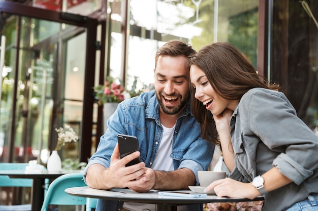 Aantrekkelijk jong koppel dat verliefd is op de lunch terwijl ze buiten aan de cafétafel zitten, met behulp van mobiele telefoon