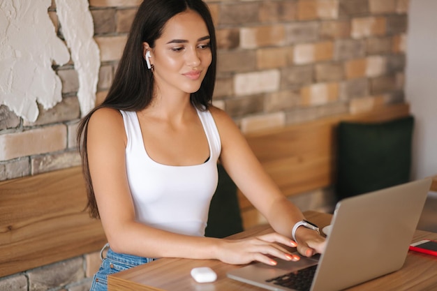 Aantrekkelijk jong meisje gebruikt draadloze hoofdtelefoons en werkt aan laptop vrouw in wit t-shirt en denim