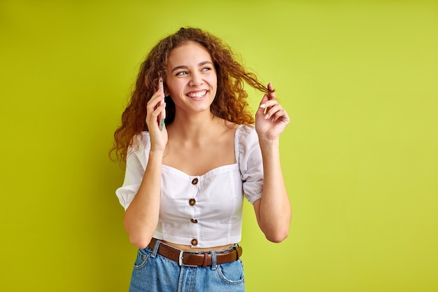 Aantrekkelijk jong meisje in blouse praten aan de telefoon met vriendin, gekrulde vrouw geniet van het delen van nieuws met iemand, geïsoleerd over groene ruimte in de studio