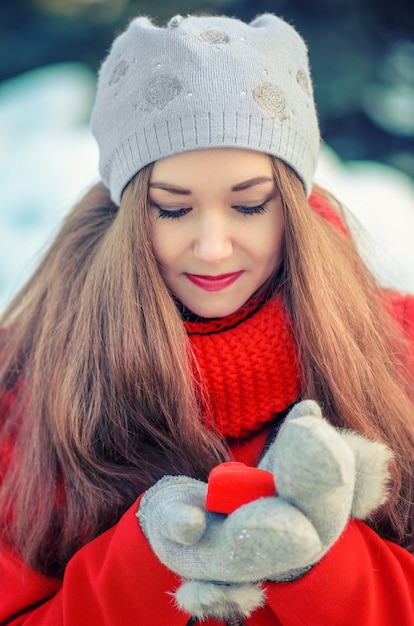 Aantrekkelijk jong meisje met lang haar in een rode jas en sjaal met het geschenk in een doos in de vorm van een hart in handen.