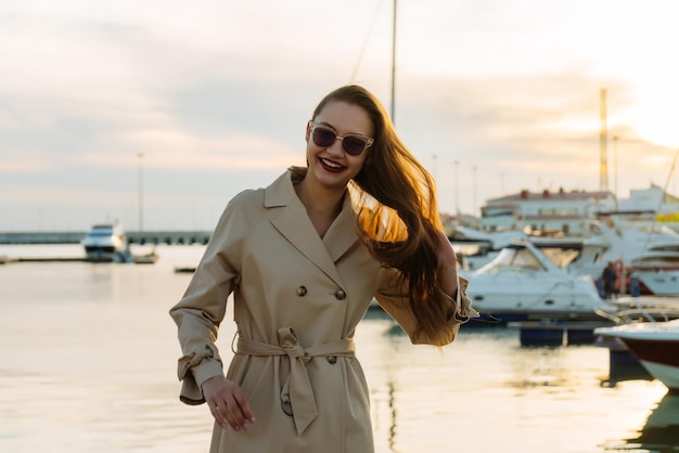 Aantrekkelijk langharig meisje in zonnebril en een beige jas staat 's avonds in de zeehaven en glimlacht