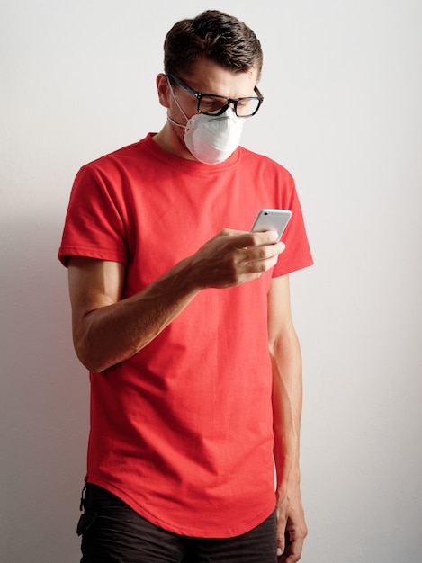 Aantrekkelijk mannetje in beschermend gezichtsmasker dat bril en rood t-shirt draagt, die mobiele telefoon houdt.