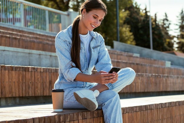 Aantrekkelijk meisje in denim kleding zit op een bankje in het park en schrijft berichten. Mooie donkerbruine vrouw