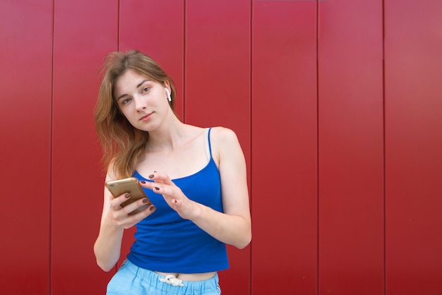 Aantrekkelijk meisje in een draadloze headset en een smartphone in zijn handen staat op de achtergrond van een rode muur en kijkt naar de camera.