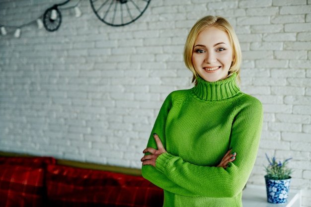 Aantrekkelijk meisje met licht haar die groene sweater dragen die zich bij witte muurmuur bevinden en, portret glimlachen.