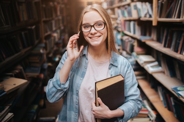 aantrekkelijk meisje permanent met een boek in haar linkerhand en praten over de telefoon. Ze kijkt recht vooruit en glimlacht. Deze vrouw is erg aardig.