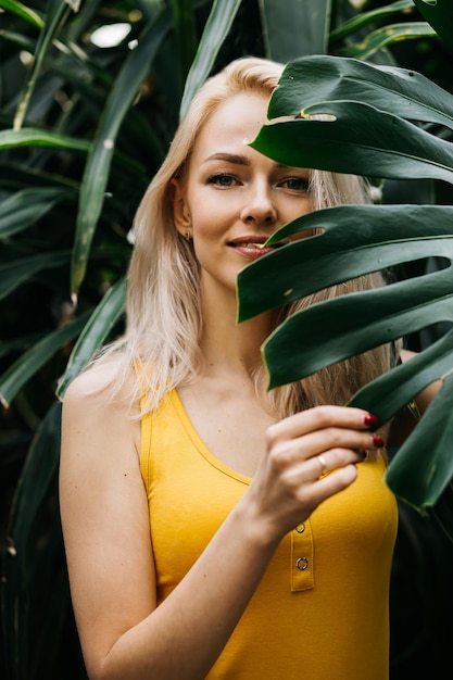 Aantrekkelijk meisje poseren in regenwoud. Close-upportret van jonge Kaukasische vrouw met blondehaar, mooie ogen die uit tropisch blad naar camera kijken. Zomer concept.