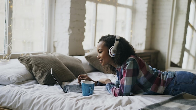 Aantrekkelijk meisje van gemengd ras luistert naar muziek tijdens het surfen op sociale media op een laptop die op bed ligt