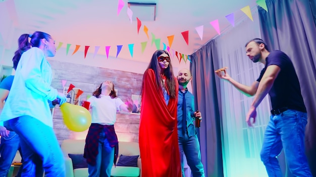 Foto aantrekkelijk meisje vol opwinding met een rode superheldenpet terwijl ze danst met haar vrienden op een wild collegefeest met neonlichten.
