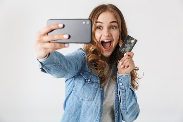 Aantrekkelijk opgewonden jong meisje dat geïsoleerd over wit staat, een selfie neemt en een plastic creditcard toont