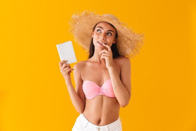 Aantrekkelijk peinzend jong meisje dat een bikini draagt die geïsoleerd over een gele muur staat, met paspoort