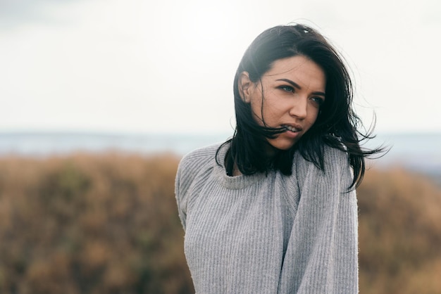 Aantrekkelijk portret van brunette blanke jonge vrouw met winderig haar die buiten droomt tegen de achtergrond van de natuur Dromende vrouw met grijze trui die opzij kijkt met waaiend haar Lifestyle reizen