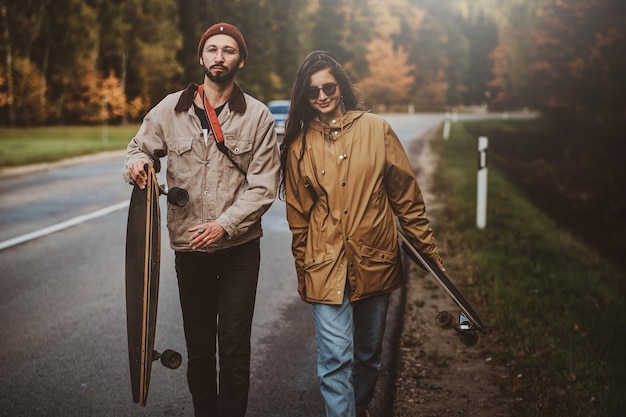 Aantrekkelijk romantisch stel loopt op de weg omringd door herfstbomen terwijl ze hun longboards vasthouden.