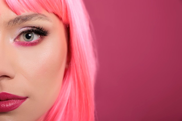 Aantrekkelijk schoonheidsmodel dat de helft van het gezicht op de camera in de studio toont, met roze haar en make-upcosmetica. Stijlvolle schattige vrouw die zich sensueel en zorgeloos voelt, plezier heeft op de achtergrond. Detailopname.