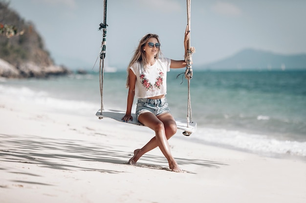 Aantrekkelijk slank meisje op het touw schommels, zandstrand tegen de blauwe baai, zonnige dag, ontspanning.