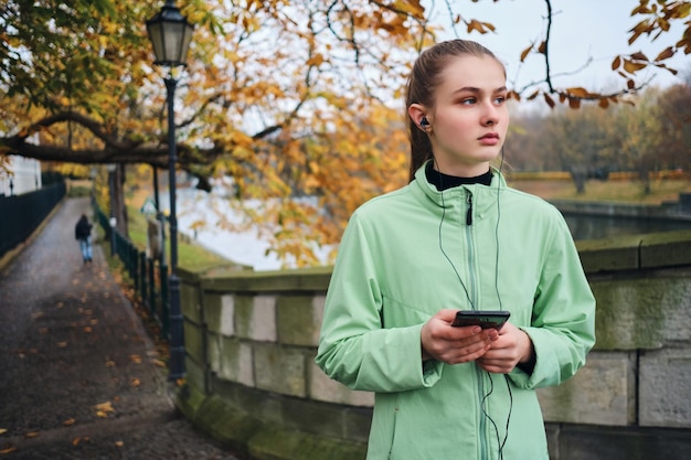 Aantrekkelijk sportief meisje in oortelefoons met mobiele telefoon die na het rennen in het herfststadspark loopt